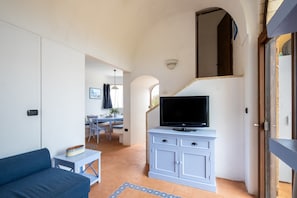Een woonkamer met een bank, een televisie op een kast, een eethoek achterin en een trap die naar een ander niveau leidt.