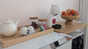 A kitchenette with a white refrigerator, a white kettle, a teapot, a jar, a bowl of fruit, and a coffee machine. - La loge Diane à deux pas du château (Chantilly)