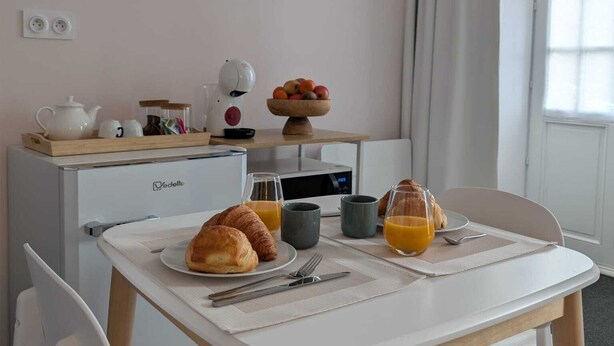 A breakfast setup with croissants, orange juice, and a fruit bowl on a white table in a room with a kitchenette, a window with curtains, and wall decorations. - La loge Diane à deux pas du château (Chantilly)