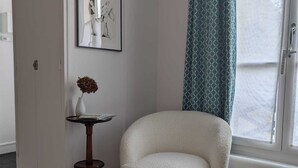 A corner room with a white chair, a small table with a vase, a framed picture, and a patterned rug. - La loge Diane à deux pas du château (Chantilly)