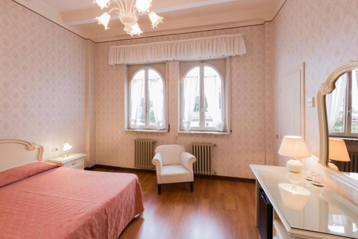 A bedroom with a bed, a chair, a dresser, a mirror, and two windows with a view of buildings outside.