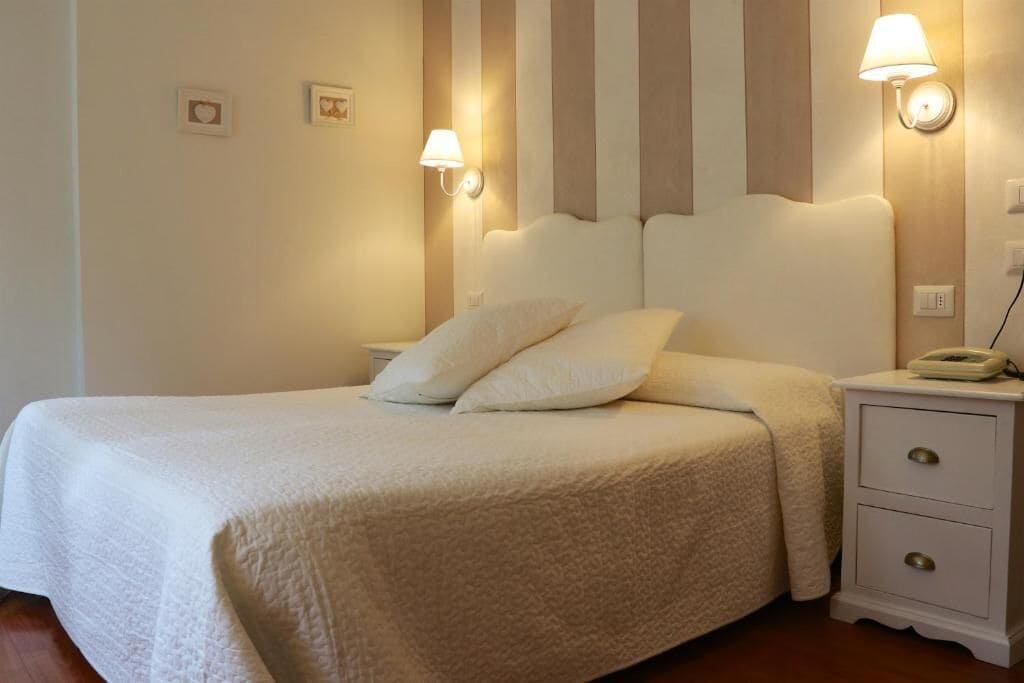 A neatly made bed with a white comforter and pillows, a bedside table with a lamp, and a wall-mounted light fixture.