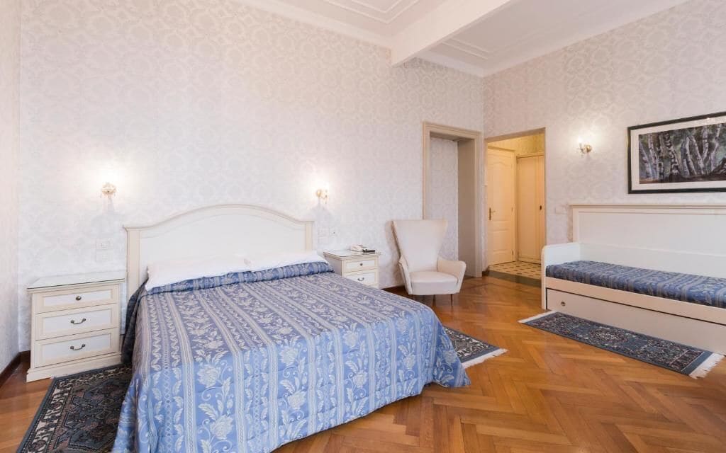 A bedroom with a large bed, two bedside tables, a chair, a smaller bed, and a painting on the wall.