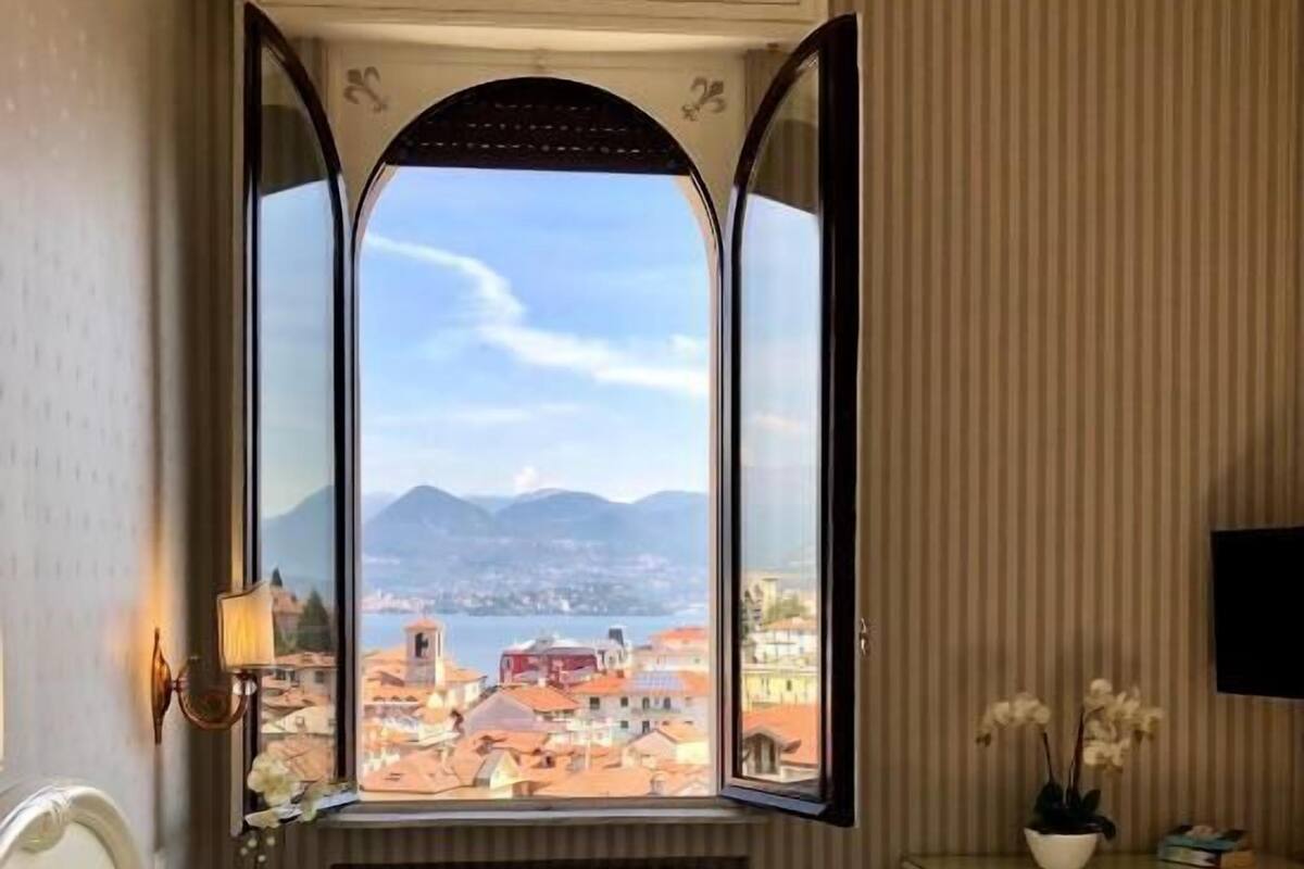 A hotel room with a bed, a desk, a radiator, a window with a view of a city and mountains, and a wall-mounted TV.
