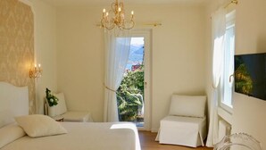 A bedroom with a bed, two bedside tables, a chair, a chandelier, a window with curtains, and a painting on the wall. - Hotel du Parc (Stresa)
