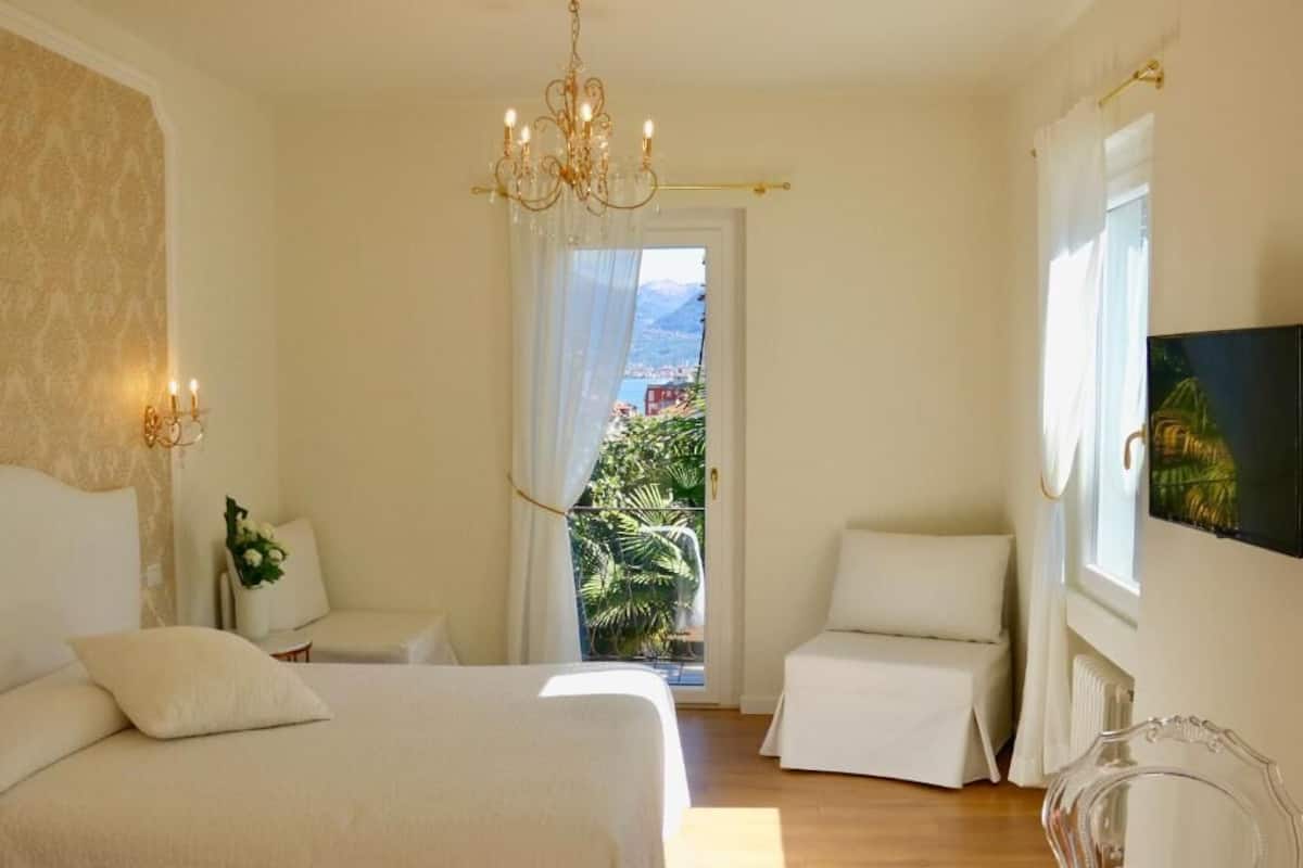 A bedroom with a bed, two bedside tables, a chair, a chandelier, a window with curtains, and a painting on the wall.