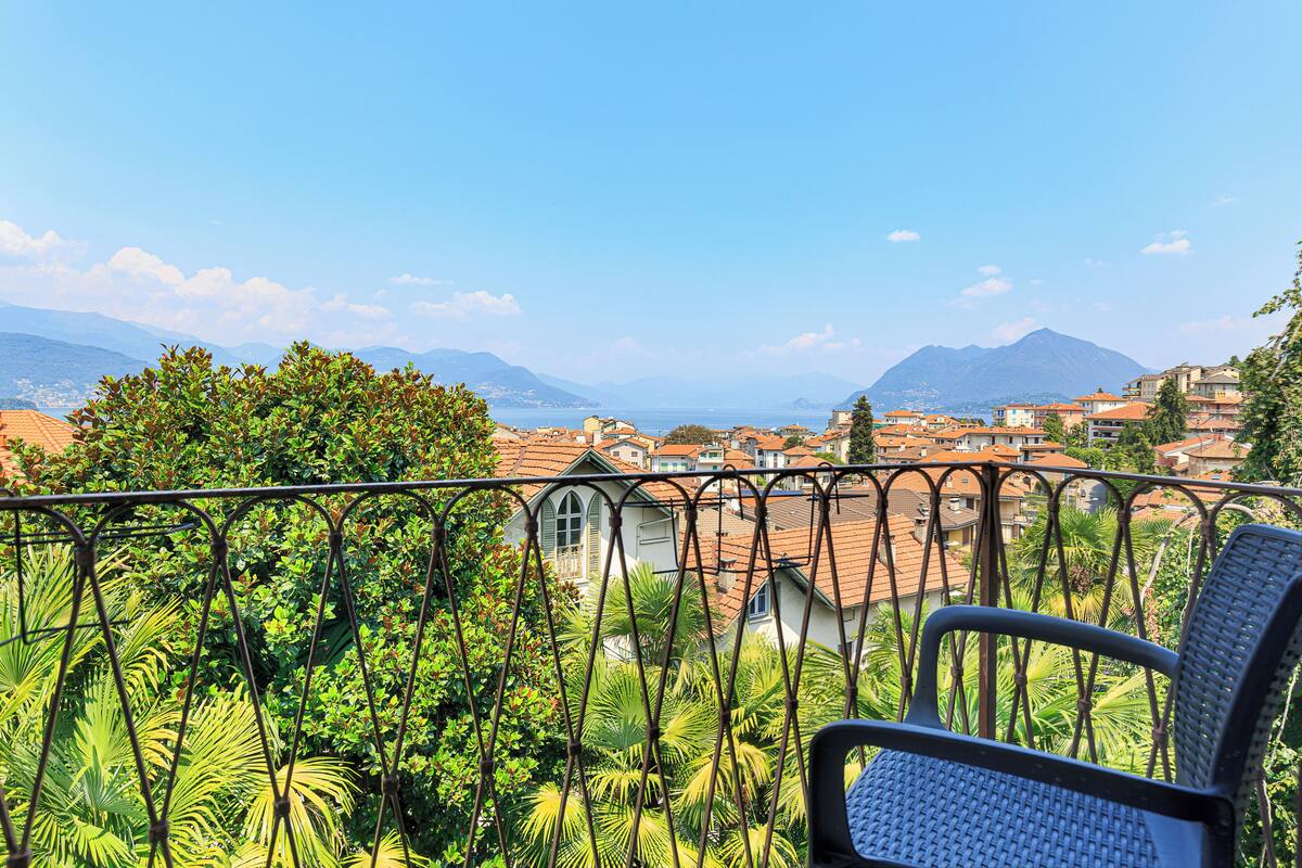A balcony with a view of a town, a chair, and lush greenery.