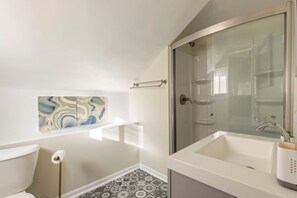 A bathroom with a glass shower enclosure, a white sink, a toilet, and a marble-patterned shelf. - South Euclid Joy - 5BR Home (South Euclid)