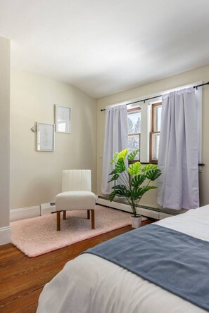 A bedroom with a bed, a chair, a radiator, a window with curtains, and a plant. - South Euclid Joy - 5BR Home (South Euclid)