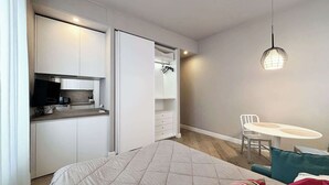 Une chambre compacte comprenant un lit, une petite kitchenette, une table à manger avec une chaise et un luminaire suspendu.