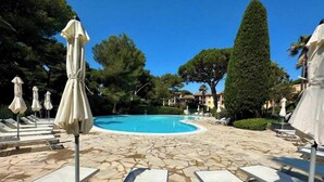 Uma piscina com espreguiçadeiras e guarda-sóis, cercada por árvores e edifícios.