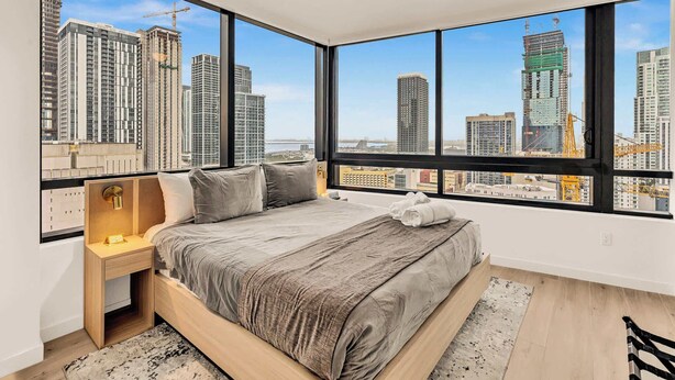 Une chambre à coucher avec un grand lit, une table de chevet, une lampe et une vue sur les immeubles de la ville, visible à travers de grandes fenêtres.