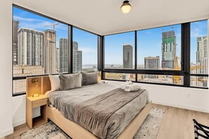 Un dormitorio con una cama grande, una mesita de noche, una lámpara y vistas a edificios de la ciudad a través de amplias ventanas.