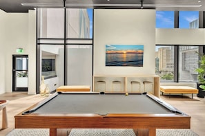 Una mesa de billar en una habitación moderna con ventanas amplias, un cuadro con atardecer y una zona de estar con vista a la ciudad.