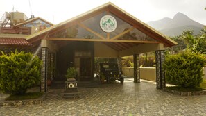 A building with a sign reading 'Hill Down Home Stay,' a covered entrance, a vehicle, and a mountainous backdrop. - Hill Down Homestay (Chikkamagaluru)
