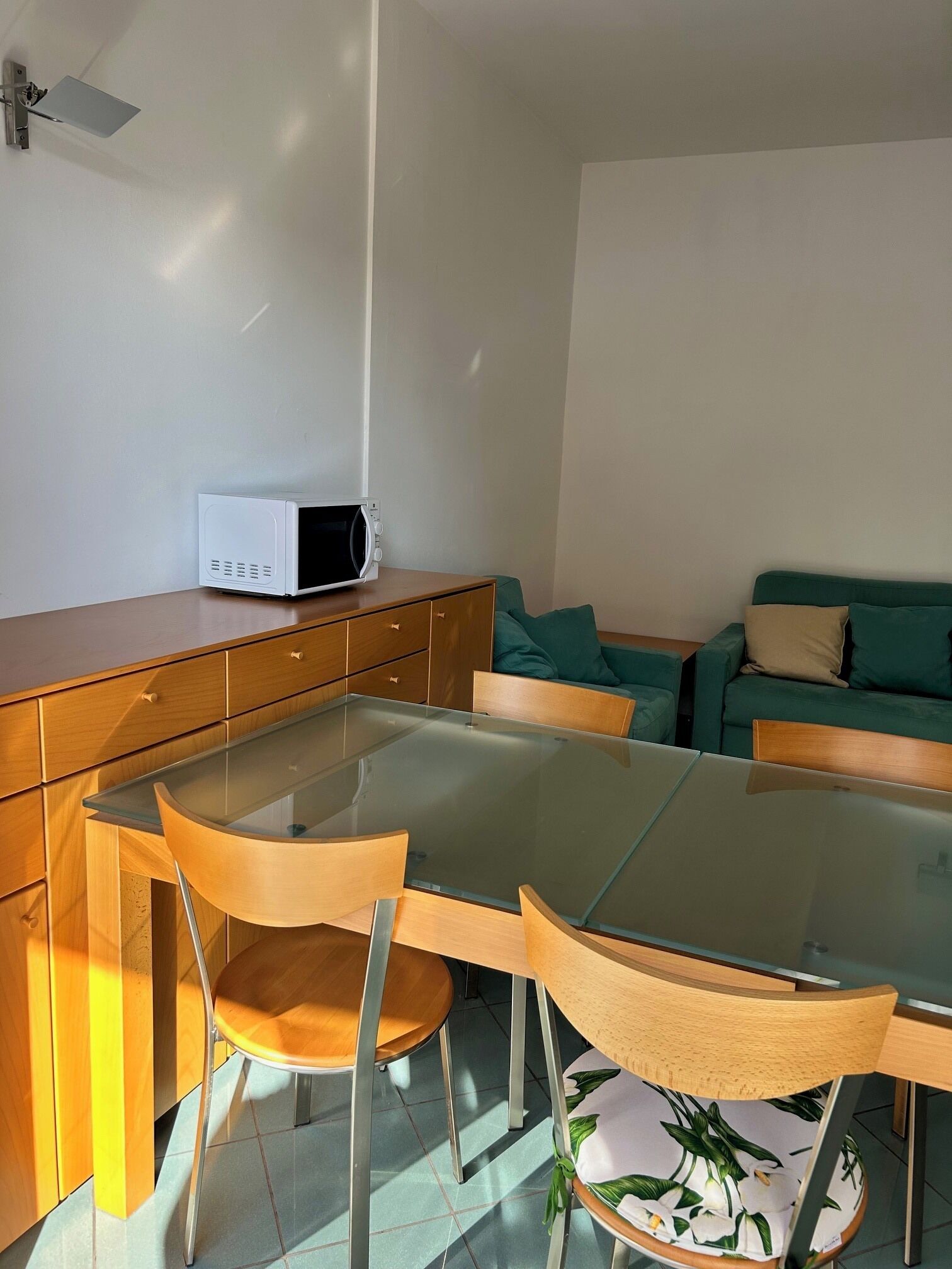 A modern dining area with a glass table, wooden chairs, a microwave on a wooden cabinet, and a teal sofa with cushions.