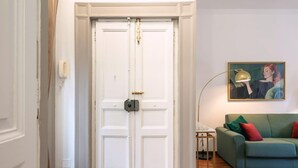 A closed white door with a security lock, a green sofa with red cushions, a small table with a lamp, and a framed picture on the wall. - Luxury Apartment for 5 with Terrace in Chiaia (Napoli)