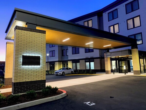 A modern hotel entrance with a covered walkway, a car parked in the lot, and a clear sky.