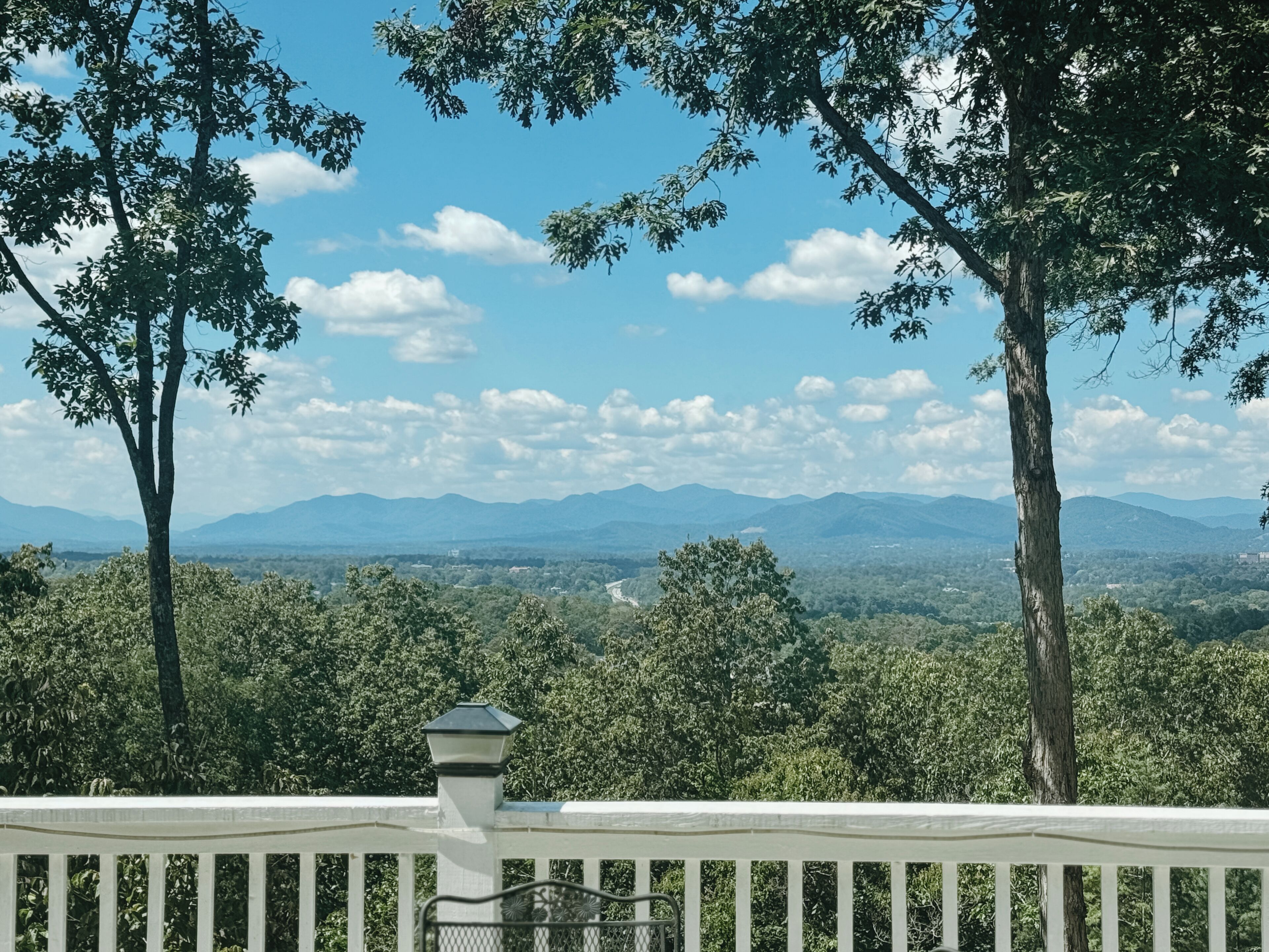 A view from a balcony with a railing, overlooking a forested landscape and distant mountains under a clear sky.