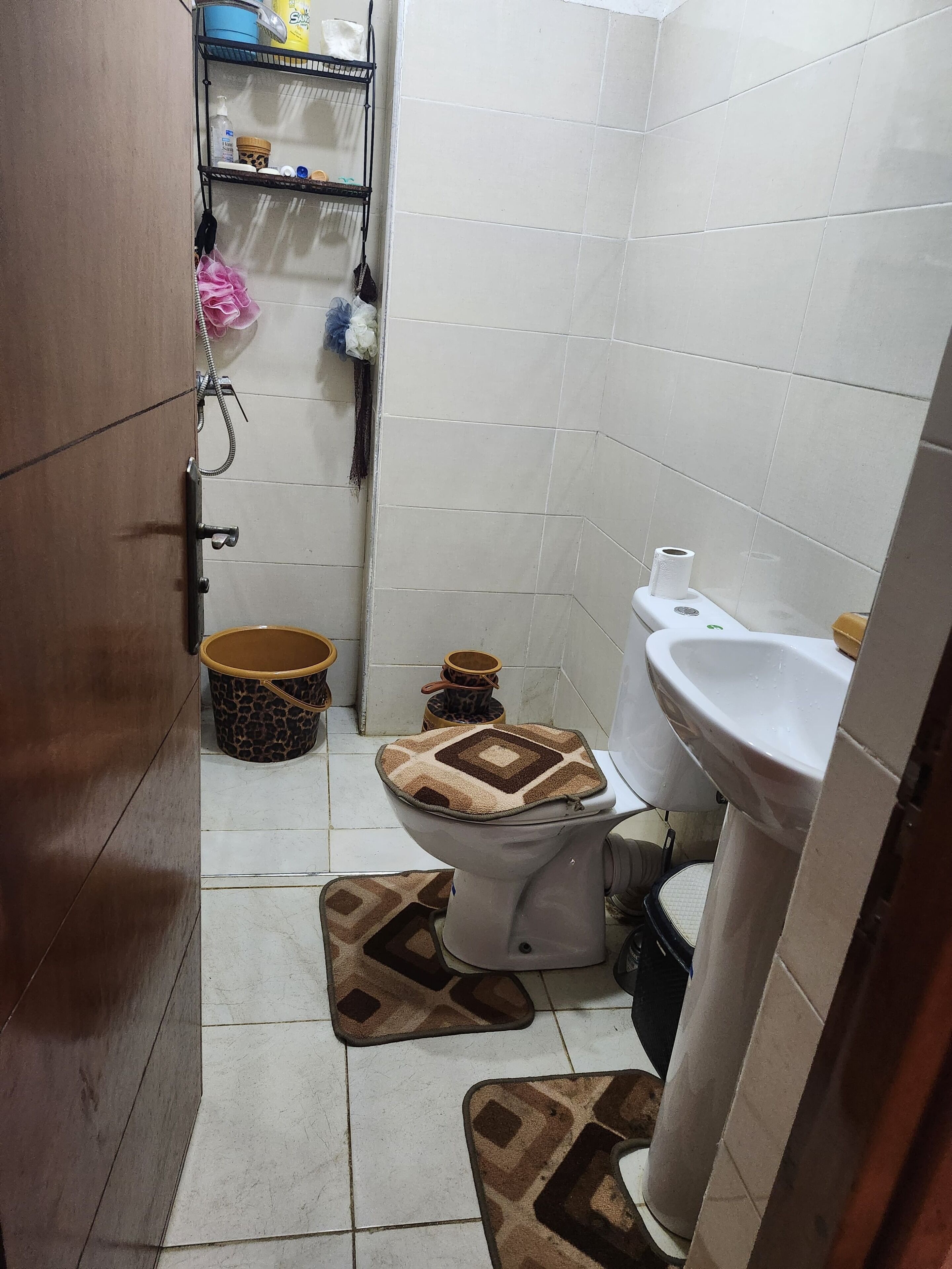 A small bathroom with a toilet, a sink, a shower, and a shelf with toiletries.