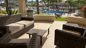 A covered outdoor seating area with wicker furniture, a pool, and palm trees in the background. - Beautifully Maintained 2-bed Condo in Las Mañanitas (San José del Cabo)