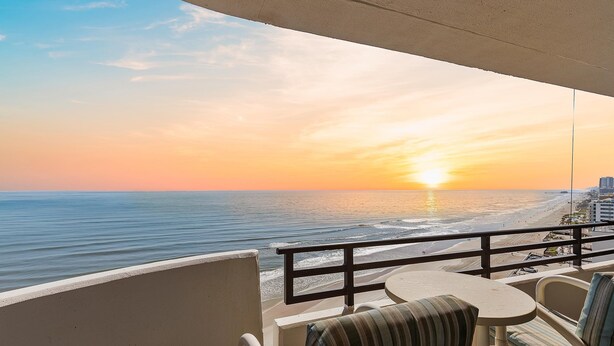 Ein Balkon mit Blick auf das Meer bei Sonnenuntergang, ein Tisch und zwei Stühle.