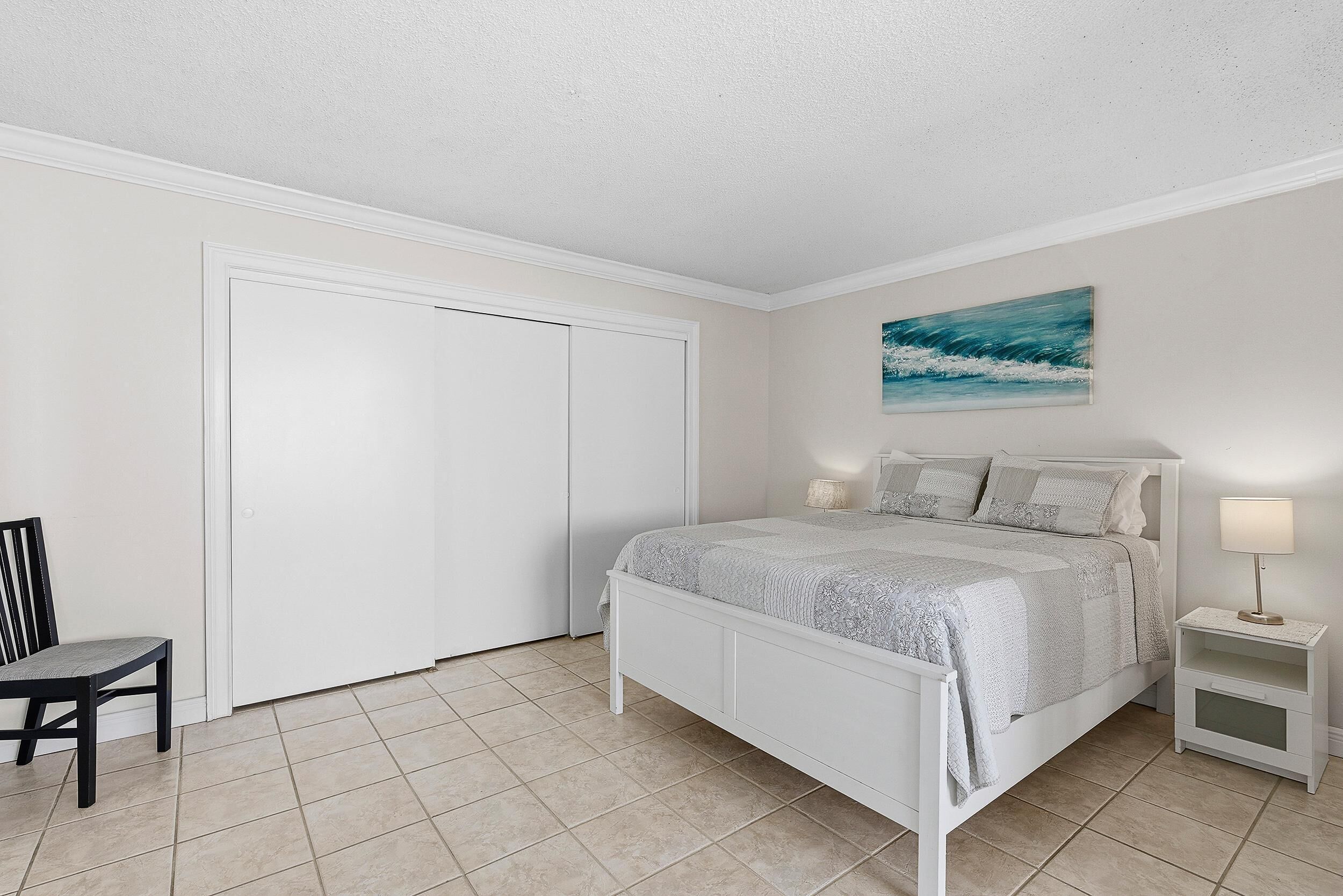 A bedroom with a bed, a chair, a nightstand, a painting, and a door.