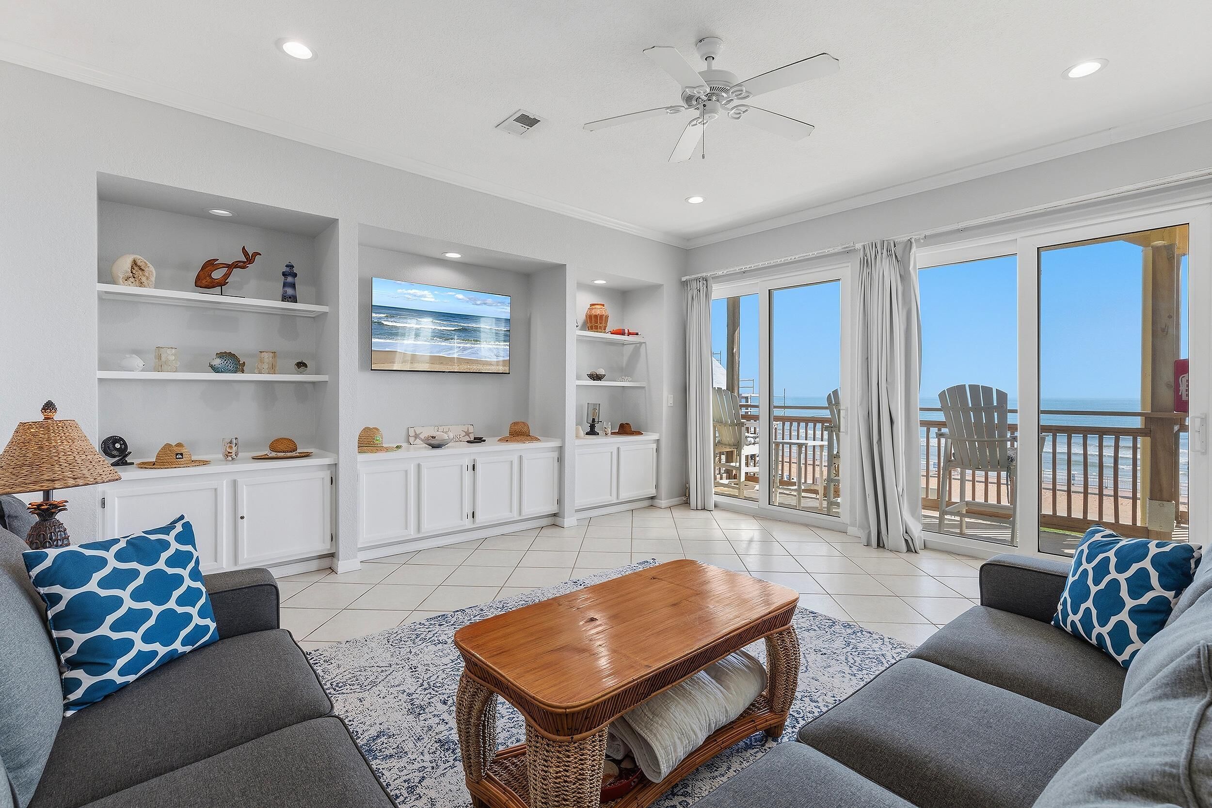 A spacious living area with a large window overlooking the beach, a ceiling fan, and a view of the ocean.
