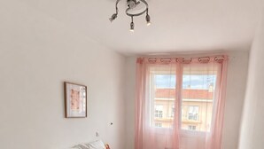 Ein Schlafzimmer mit einem Bett, einem Nachttisch, einer Lampe, einem rosa Teppich und einem Fenster mit Vorhängen.