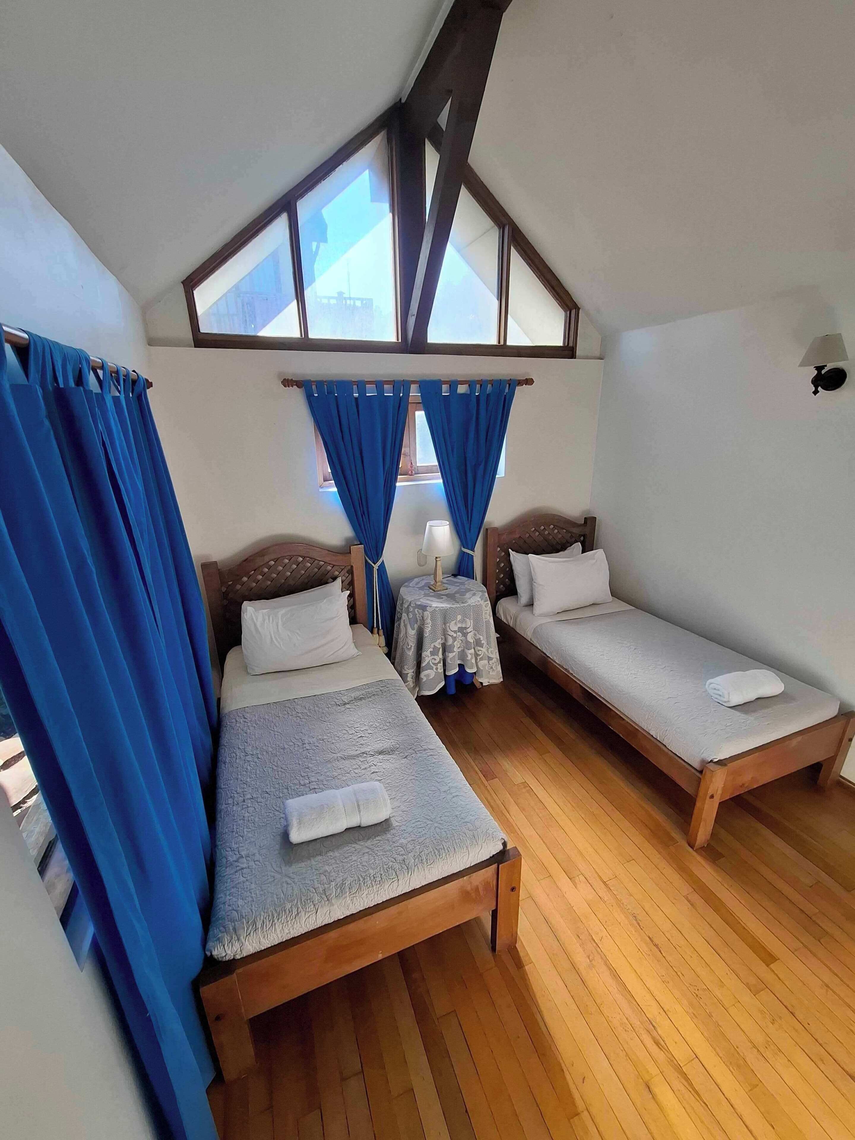 Ein Zimmer mit zwei Holzbetten, einem kleinen Tisch mit Lampe und einem Fenster mit blauen Vorhängen.