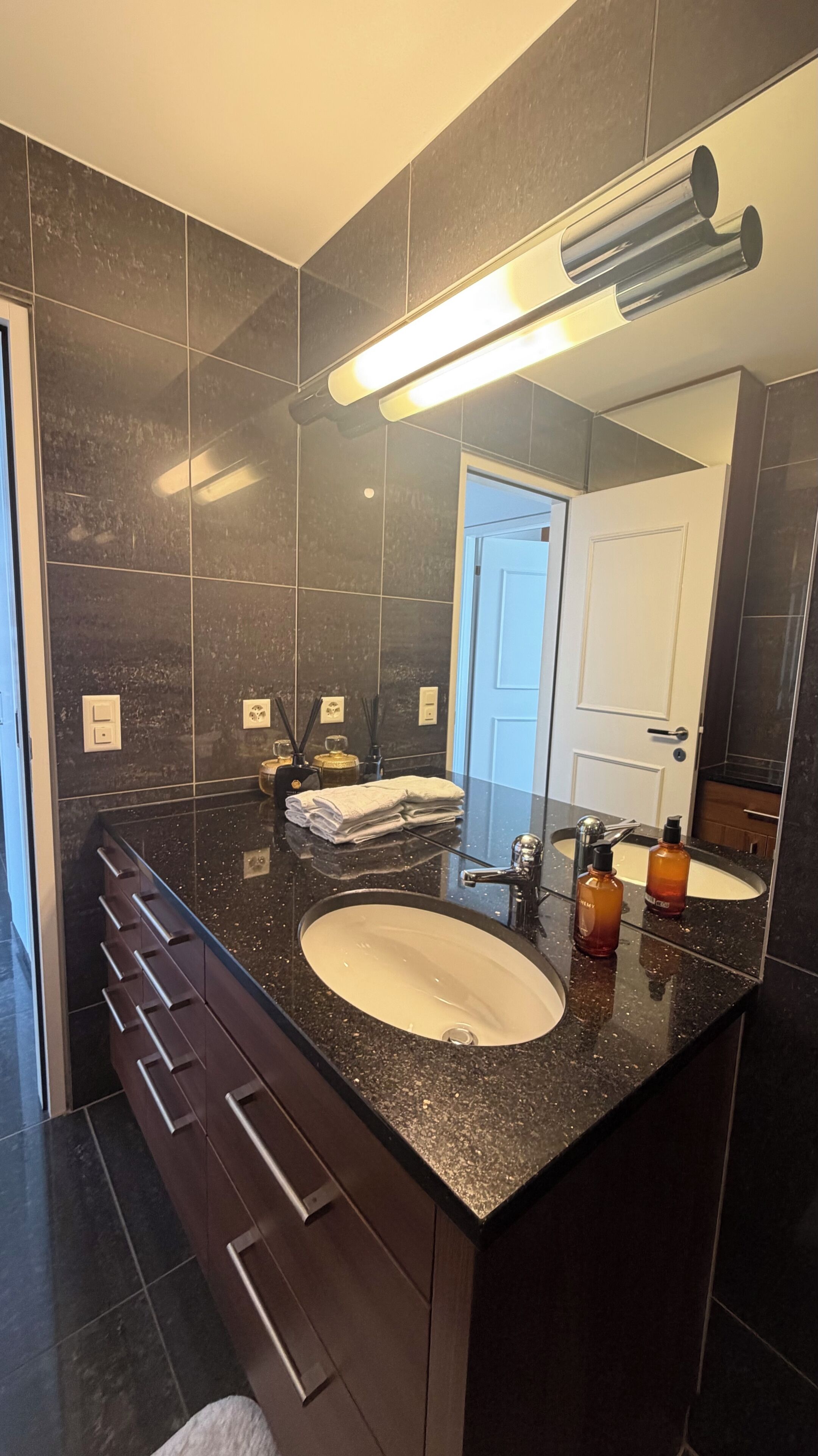 Un baño moderno con tocador de mármol oscuro, un espejo grande e lavamanos doble.