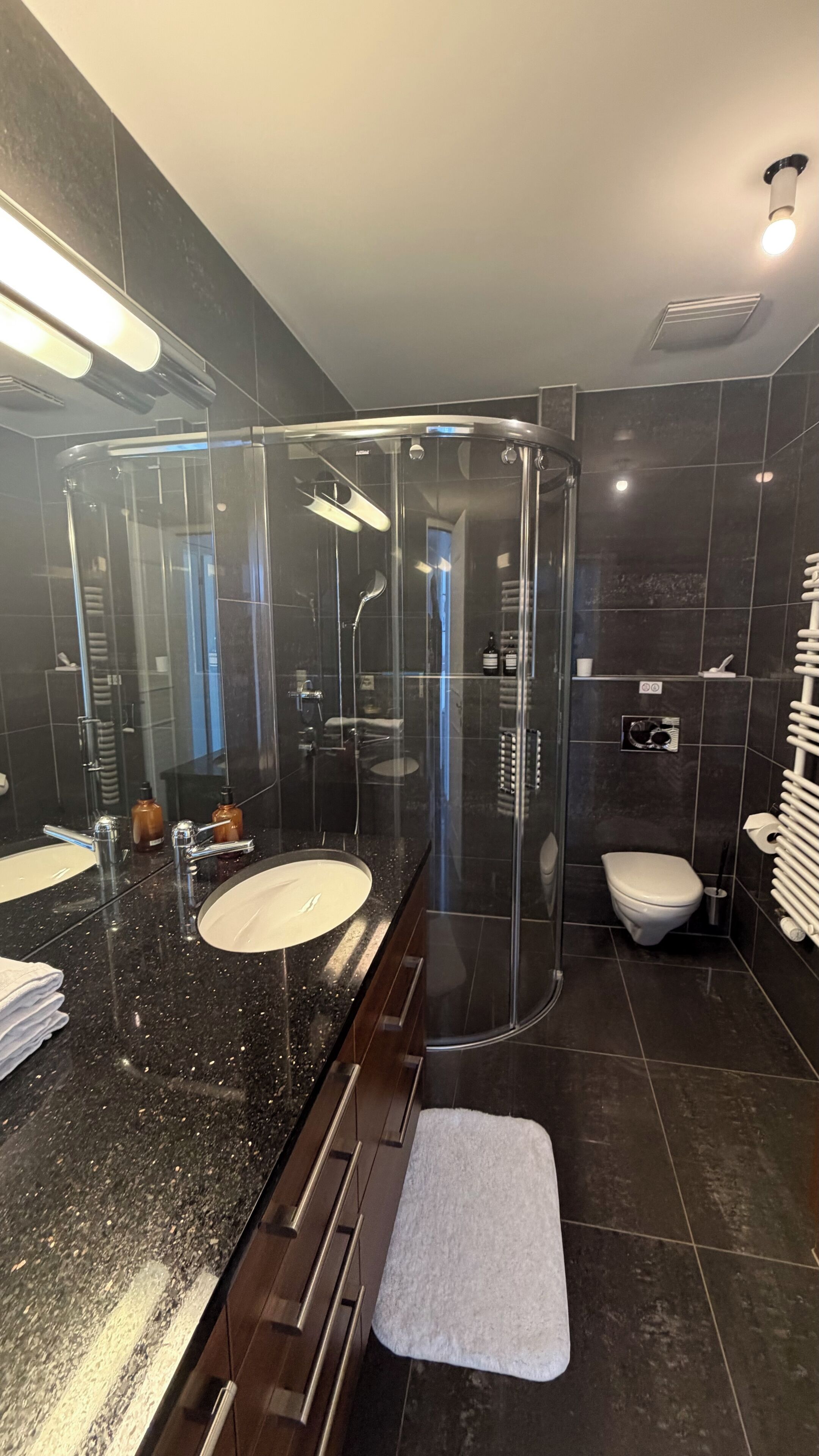 A modern bathroom with a glass-enclosed shower, a large mirror, and a dark tiled floor.