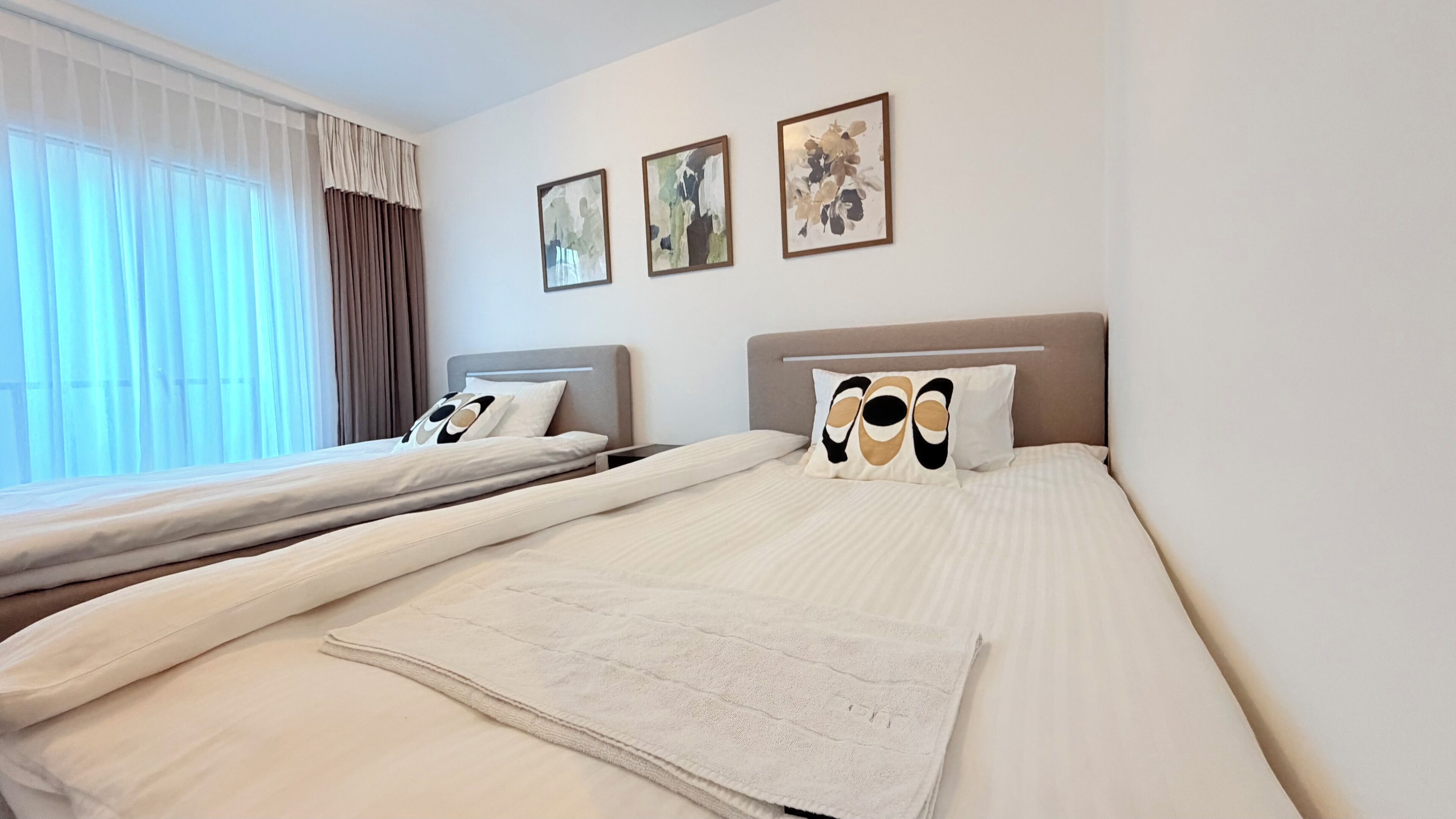 Una habitación de hotel con dos camas, un ventanal con cortinas y tres cuadros enmarcados en la pared.
