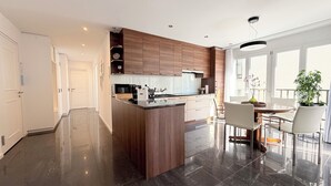 A modern kitchen with a central island, wooden cabinets, and a dining area with a glass table and chairs. - Center Park Apartment (Interlaken)
