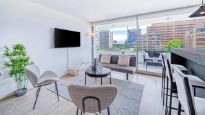 En moderne stue med et fladskærms-TV, en sofa, et spiseområde og et stort vindue med udsigt over byen.