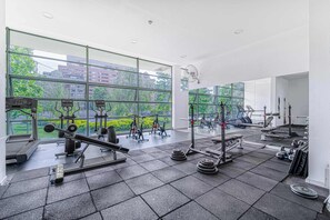 A gym with various exercise equipment, large windows, and a view of buildings and trees. - Nordic Home - 2BR, Parking, Wifi & Shopping Mall (Santiago)