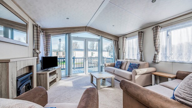 A modern living room with a flat-screen TV, beige sofas, a coffee table, and large windows. - Lovely Lakeside Holiday Park Home  - Pass the Keys (Chichester)