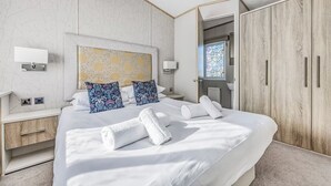 A modern hotel room with a large bed, two bedside tables, a wardrobe, and a bathroom visible through an open door. - Lovely Lakeside Holiday Park Home  - Pass the Keys (Chichester)