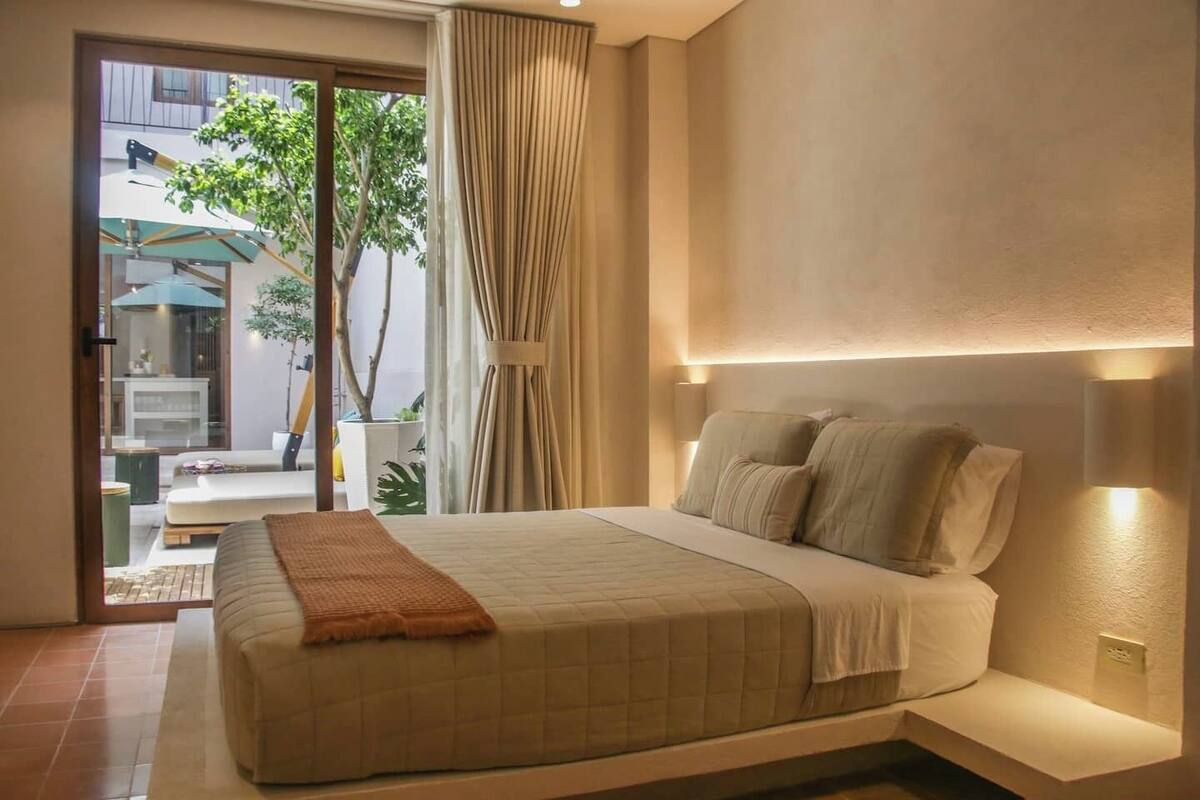 Une chambre à coucher avec un lit, une table de chevet, une fenêtre avec des rideaux et une vue sur l’extérieur.