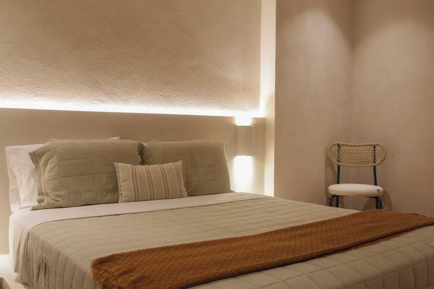 A bed with a headboard, pillows, a blanket, and a chair in a room with a plain wall and a light fixture above the bed.