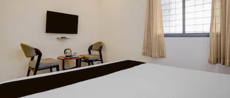 A hotel room with a bed, two chairs, a small table with a coffee pot, and a TV on the wall.