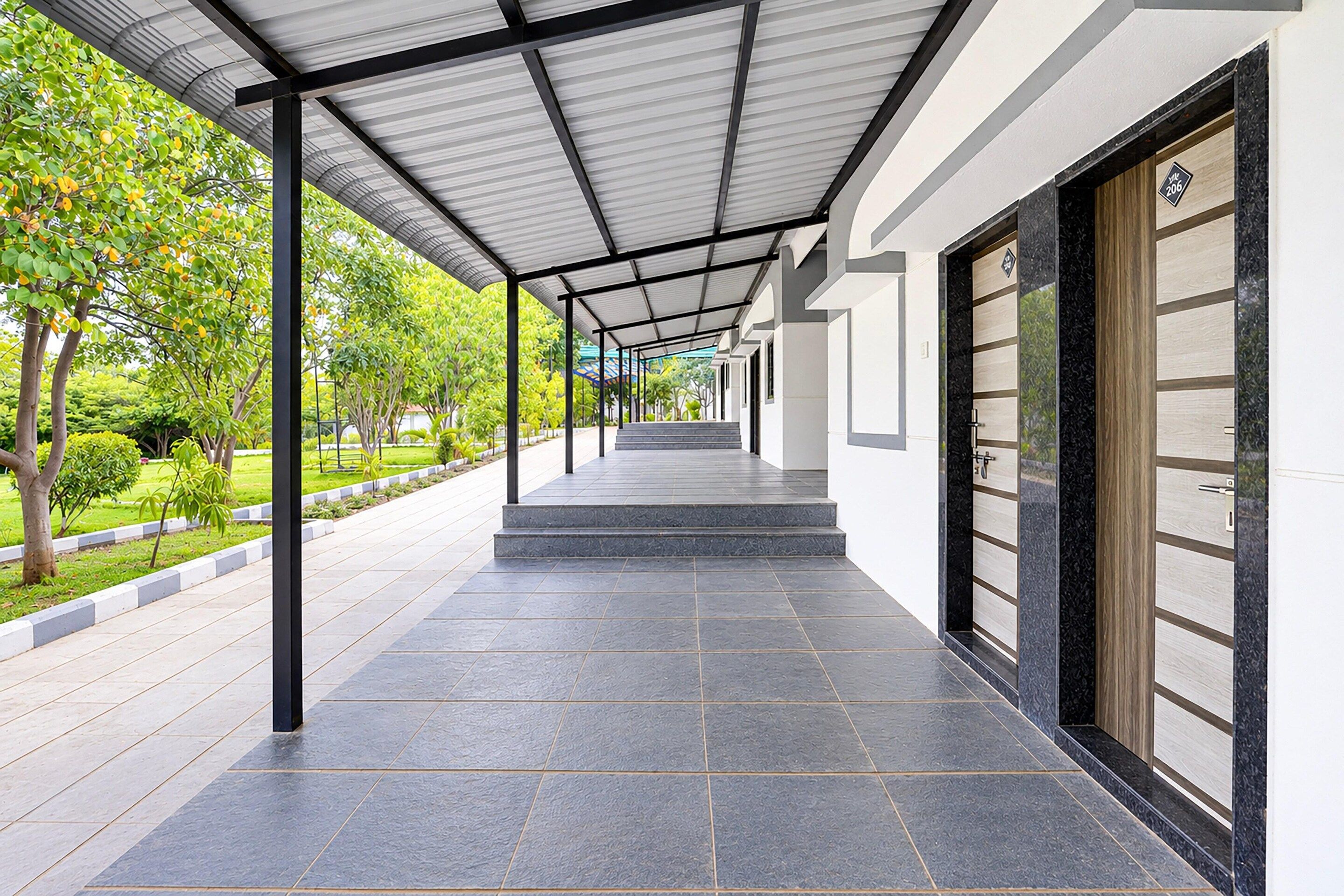 A covered walkway with a tiled floor, flanked by greenery and a building with a large door.