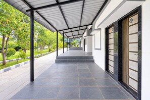 A covered walkway with a tiled floor, flanked by greenery and a building with a large door.