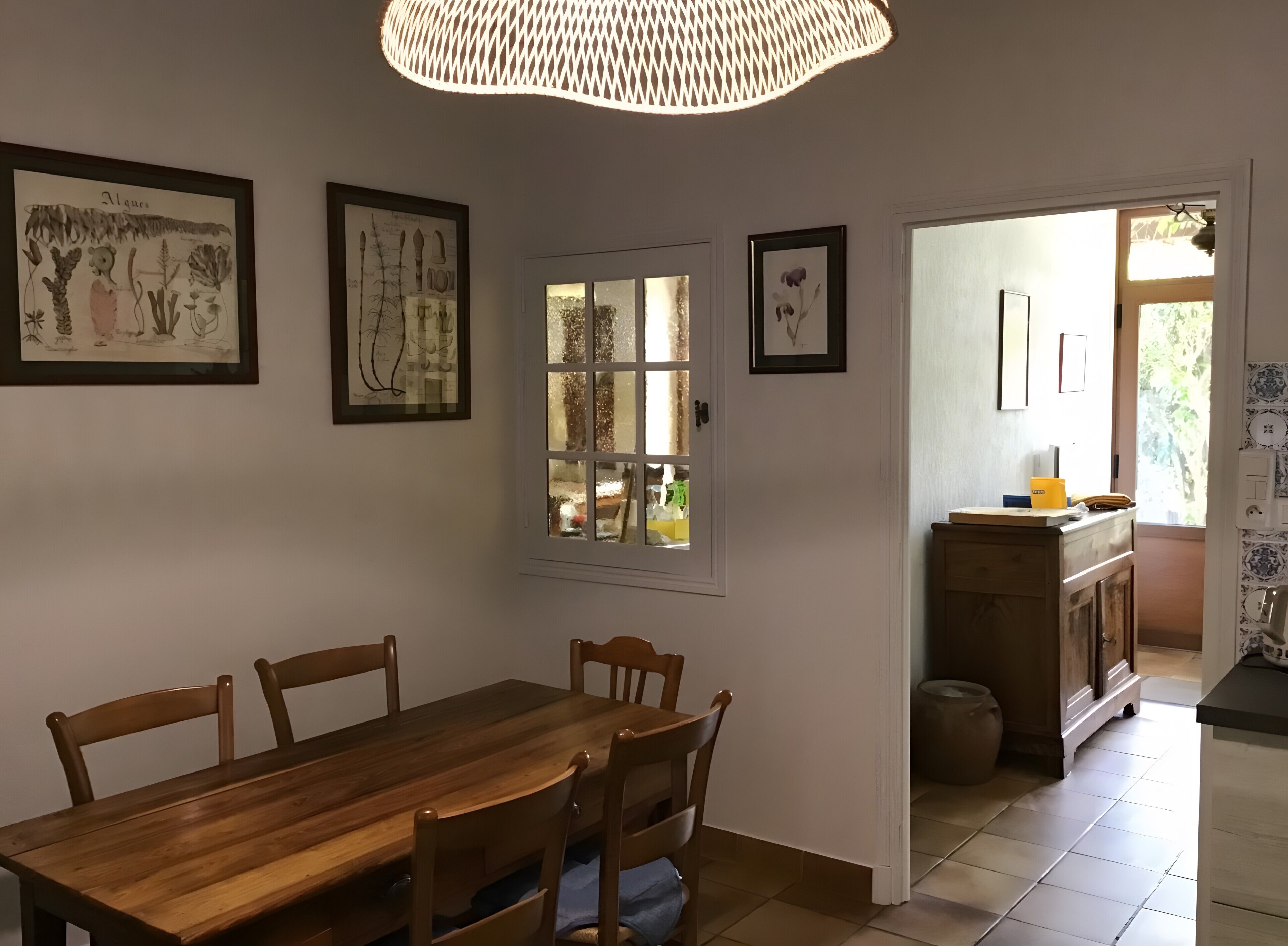 A dining area with a wooden table and chairs, framed botanical prints on the wall, a window with a view, and a kitchen area with a counter and cabinets.