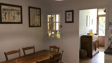 A dining area with a wooden table and chairs, framed botanical prints on the wall, a window with a view, and a kitchen area with a counter and cabinets.