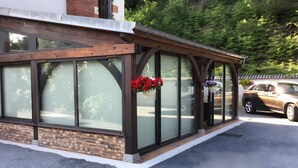 A building with large glass windows, a brick lower section, and a covered porch area with hanging flower baskets. - Holiday Home "Gîte for 6 People" with Private Terrace and Wi-Fi (La Chapelle)