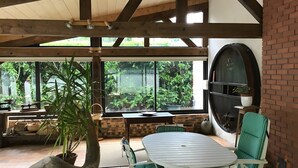 An outdoor patio with a table and chairs, a large potted plant, and a view of greenery through large windows. - Holiday Home "Gîte for 6 People" with Private Terrace and Wi-Fi (La Chapelle)