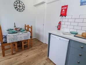 A kitchen with a dining table set for two, a clock on the wall, a fire extinguisher, and a dishwasher. - Dolphin Cottage (Weymouth)
