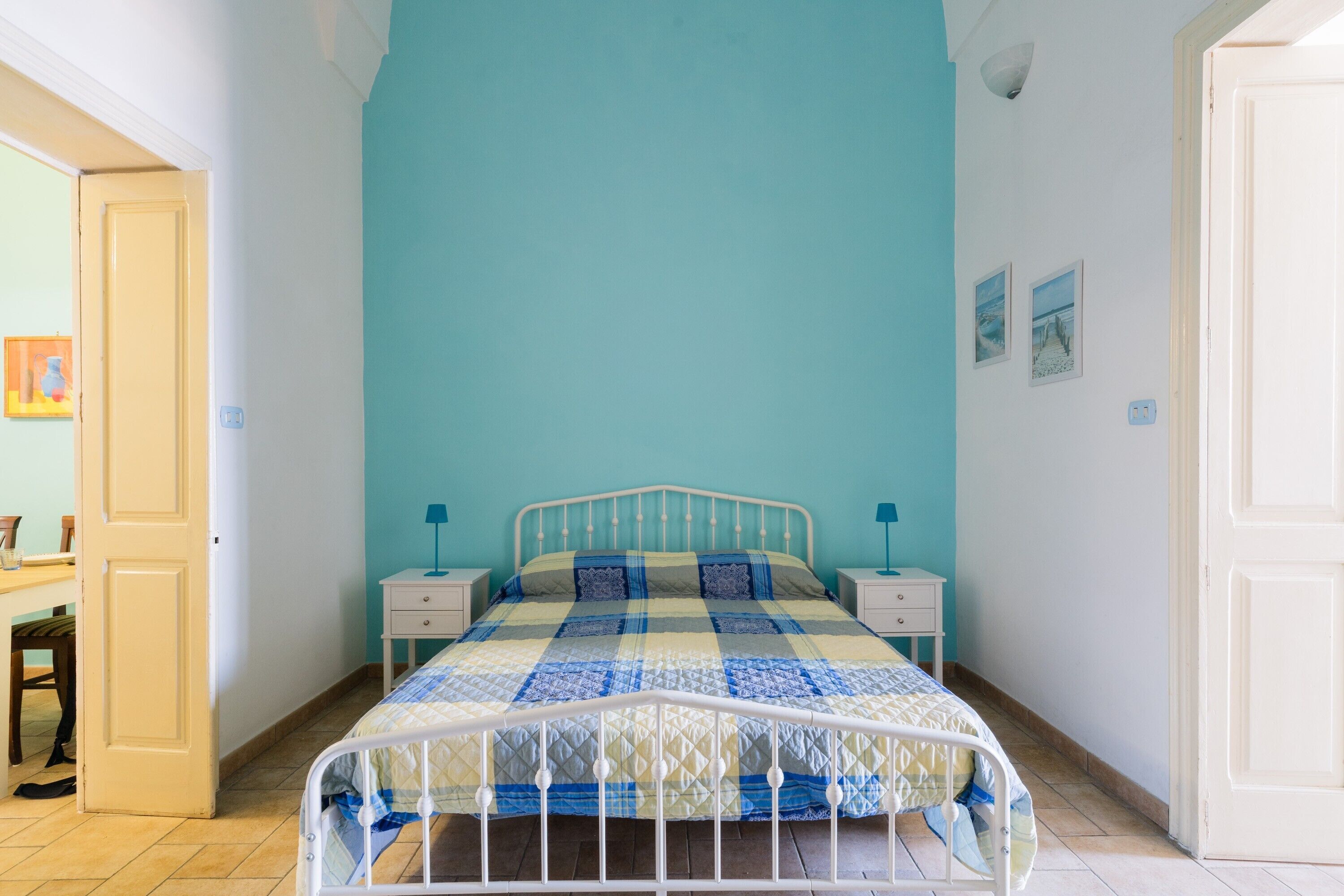 A bedroom with a bed, two bedside tables, a nightstand, a door, and a wall with two pictures.