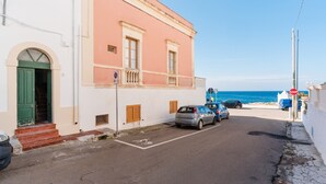 Eine schmale Straße mit parkenden Autos, ein Gebäude mit pinkfarbener Fassade und freier Meersicht.